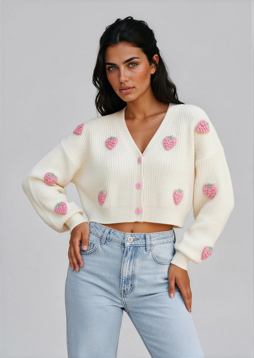 Woman wearing a white cardigan with strawberry patterns against a stone wall.